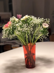 A bouquet of small flowers with delicate white blooms and a few pink and purple flowers in a red transparent vase placed on a white tabletop. The flowers are arranged with leafy green stems, creating a fresh and lively appearance.