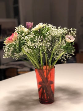 A bouquet of small flowers with delicate white blooms and a few pink and purple flowers in a red transparent vase placed on a white tabletop. The flowers are arranged with leafy green stems, creating a fresh and lively appearance.