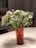 A bouquet of small flowers with delicate white blooms and a few pink and purple flowers in a red transparent vase placed on a white tabletop. The flowers are arranged with leafy green stems, creating a fresh and lively appearance.