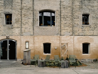 A rustic brick building with arched windows and doors, featuring a subtle beige and gray color scheme. Wooden barrels and green metal chairs are arranged in front of the building, suggesting an outdoor seating area. The building has the number 93 and a small sign displaying 'Beer Barr'. The overall setting appears casual and inviting.
