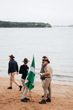 A group gathered on a sunlit beach, engaged in a lively discussion with hiking gear and notebooks.