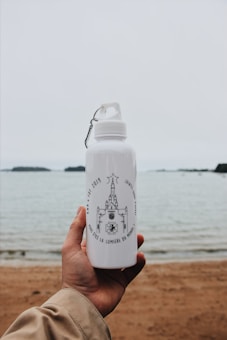 A hand holding a white metal water bottle with a design featuring a church-like structure and inscriptions in French, against a backdrop of a sandy beach and calm water, possibly a lake or ocean, with a cloudy sky overhead.
