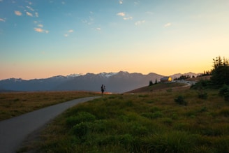 A scenic landscape capturing a person with a camera on a tripod standing on a grassy path, with rolling hills leading to a small house near the edge. In the background, a range of snow-capped mountains is visible against a colorful sky transitioning from orange to blue as the sun sets.