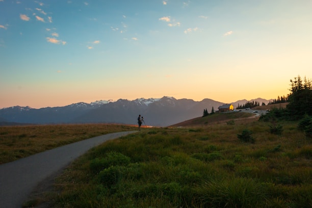 A scenic landscape capturing a person with a camera on a tripod standing on a grassy path, with rolling hills leading to a small house near the edge. In the background, a range of snow-capped mountains is visible against a colorful sky transitioning from orange to blue as the sun sets.