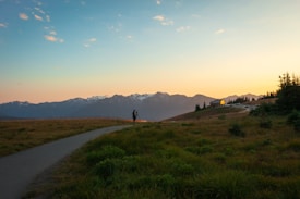 A scenic landscape capturing a person with a camera on a tripod standing on a grassy path, with rolling hills leading to a small house near the edge. In the background, a range of snow-capped mountains is visible against a colorful sky transitioning from orange to blue as the sun sets.