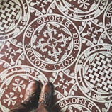 Intricate patterned floor tiles feature ornate white designs on a brown background, including floral and heraldic motifs with Latin script encircling the patterns. A pair of brown leather shoes stand on the tiles, partially visible in the image.