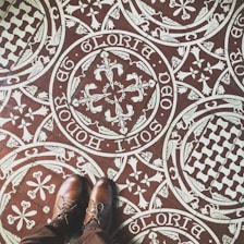 Intricate patterned floor tiles feature ornate white designs on a brown background, including floral and heraldic motifs with Latin script encircling the patterns. A pair of brown leather shoes stand on the tiles, partially visible in the image.