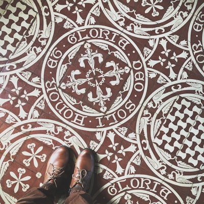 Intricate patterned floor tiles feature ornate white designs on a brown background, including floral and heraldic motifs with Latin script encircling the patterns. A pair of brown leather shoes stand on the tiles, partially visible in the image.