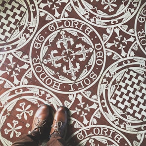 Intricate patterned floor tiles feature ornate white designs on a brown background, including floral and heraldic motifs with Latin script encircling the patterns. A pair of brown leather shoes stand on the tiles, partially visible in the image.