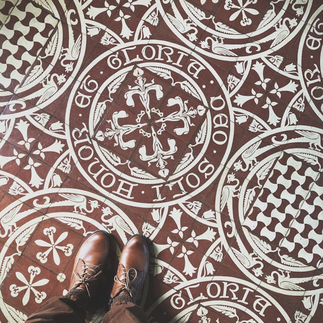 Intricate patterned floor tiles feature ornate white designs on a brown background, including floral and heraldic motifs with Latin script encircling the patterns. A pair of brown leather shoes stand on the tiles, partially visible in the image.
