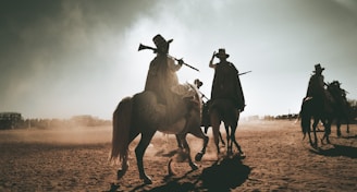 Riders on horseback are silhouetted against a dusty sky, each holding a rifle upwards. The scene is set in a barren, sandy area with distant figures and possibly a structure visible on the horizon. Shadows stretch long across the ground, adding to the dramatic and timeless feel of the scene.