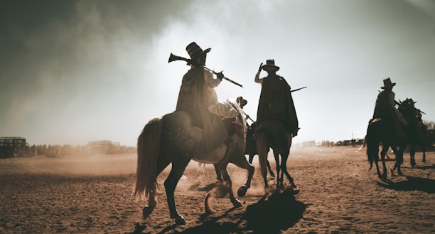 Riders on horseback are silhouetted against a dusty sky, each holding a rifle upwards. The scene is set in a barren, sandy area with distant figures and possibly a structure visible on the horizon. Shadows stretch long across the ground, adding to the dramatic and timeless feel of the scene.