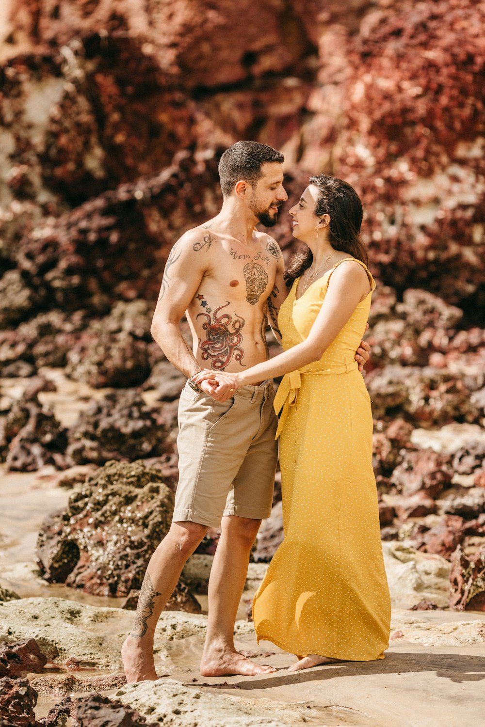 topless man hugging woman in yellow dress on seashore during daytime