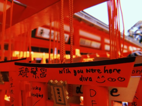 A close-up view of bright orange wooden plaques hanging from strings. The plaques contain handwritten messages, some in English and some in another language. The central plaque reads 'Wish you were here diva' alongside doodles of hearts and a smiley face. The background appears to be part of a shrine or temple with more orange architectural details.