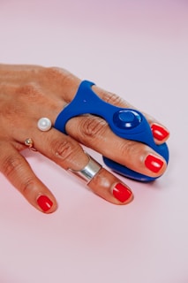 A hand with bright red nail polish is adorned with silver and pearl rings. A blue device with a button is grasped between the index and middle fingers. The background is a soft gradient of pink and white.