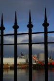 A wrought iron fence with pointed tops is prominently displayed in the foreground, casting shadows and silhouettes. In the background, an industrial scene is visible, including large silos and cranes, set against a partly cloudy sky.