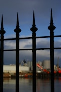 A wrought iron fence with pointed tops is prominently displayed in the foreground, casting shadows and silhouettes. In the background, an industrial scene is visible, including large silos and cranes, set against a partly cloudy sky.