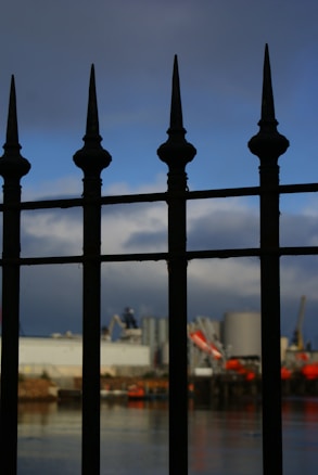A wrought iron fence with pointed tops is prominently displayed in the foreground, casting shadows and silhouettes. In the background, an industrial scene is visible, including large silos and cranes, set against a partly cloudy sky.