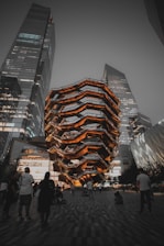 A futuristic architectural structure with layered, honeycomb-like design illuminated in warm lights stands amidst towering modern buildings. People are gathered around, some walking, others sitting on the paved ground, suggesting an urban public space.