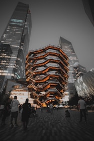 A futuristic architectural structure with layered, honeycomb-like design illuminated in warm lights stands amidst towering modern buildings. People are gathered around, some walking, others sitting on the paved ground, suggesting an urban public space.