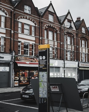 A street scene featuring a row of brick buildings with shops on the ground floor, including a candy store with a sign reading 'Sweet Portugal.' A directional street sign for West Norwood with a map is prominently displayed in the foreground. Cars are parked alongside the street.