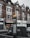 A street scene featuring a row of brick buildings with shops on the ground floor, including a candy store with a sign reading 'Sweet Portugal.' A directional street sign for West Norwood with a map is prominently displayed in the foreground. Cars are parked alongside the street.