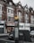 A street scene featuring a row of brick buildings with shops on the ground floor, including a candy store with a sign reading 'Sweet Portugal.' A directional street sign for West Norwood with a map is prominently displayed in the foreground. Cars are parked alongside the street.