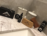 A collection of grooming products is arranged on a marble countertop. Items include a black box with grooming scissors, a tin labeled 'Beard Balm,' a bottle labeled 'Beard Oil,' a wooden brush, and a wooden comb. Decorative ceramic jars with intricate patterns stand in the background.