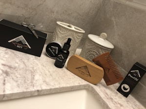 A collection of grooming products is arranged on a marble countertop. Items include a black box with grooming scissors, a tin labeled 'Beard Balm,' a bottle labeled 'Beard Oil,' a wooden brush, and a wooden comb. Decorative ceramic jars with intricate patterns stand in the background.
