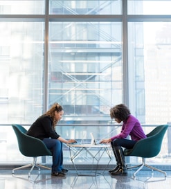 two women sits of padded chairs while using laptop computers