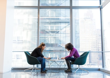 two women sits of padded chairs while using laptop computers