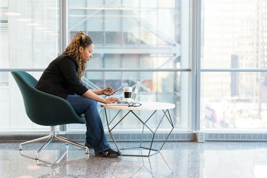 A person sits in a modern chair, focused on a laptop placed on a small circular table alongside a cup. The background features large windows with a cityscape view, suggesting a bright and professional office environment. The setting is clean and minimalistic, highlighting the individual's concentration on their work.