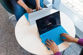 Two people are using Surface tablets with keyboards on a round, light-colored table. Their hands rest on the keyboards, suggesting they are engaged in typing or working. The setting appears to be a modern office or workspace with a speckled floor and large windows.