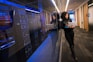 A woman is leaning against a glass wall in a hallway, holding a laptop. She is standing next to a row of server racks, with her reflection visible in the glass. The setting appears to be a modern office or data center with bright overhead lighting and a clean, professional environment.