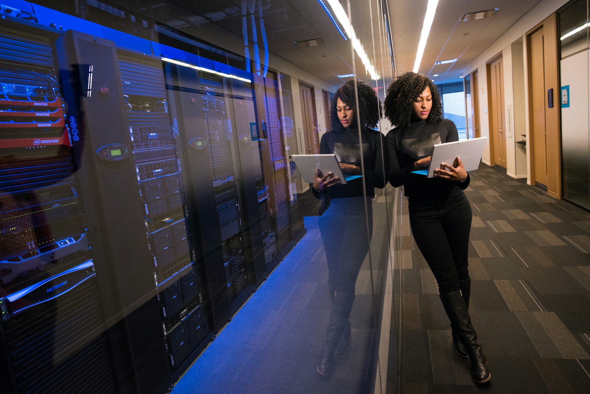 Woman leaning against a glass window looking at a laptop. On the other side of the window is an array of servers.