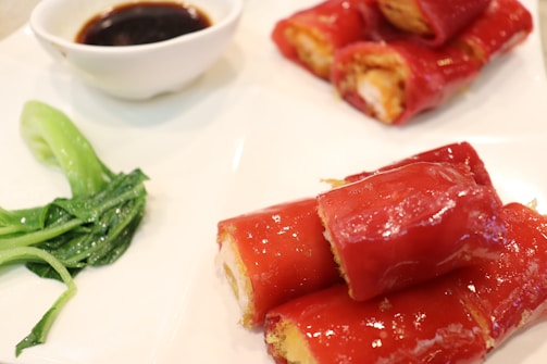 Red rice noodle rolls filled with a savory stuffing, served on a white plate. There is a small portion of green leafy vegetables and a small bowl of dark dipping sauce.