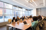 A diverse group of people are seated around a large conference table in a modern office setting. Everyone is working with laptops, which are open in front of them. Large windows in the background provide a view of neighboring buildings, and the room is well-lit with overhead lights.