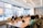 A diverse group of people are seated around a large conference table in a modern office setting. Everyone is working with laptops, which are open in front of them. Large windows in the background provide a view of neighboring buildings, and the room is well-lit with overhead lights.