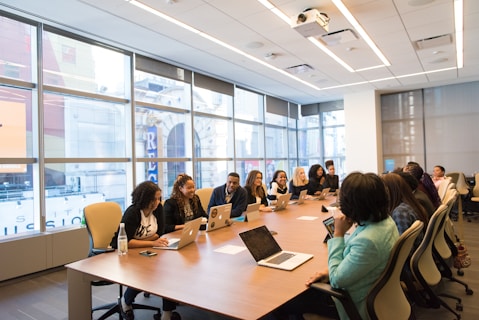 A diverse group of people are seated around a large conference table in a modern office setting. Everyone is working with laptops, which are open in front of them. Large windows in the background provide a view of neighboring buildings, and the room is well-lit with overhead lights.