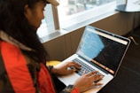 A person wearing a red jacket and cap is seated at a table, typing on a laptop displaying code. The scene appears to be in a modern office or workspace with large windows allowing natural light.