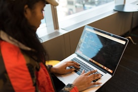 A person wearing a red jacket and cap is seated at a table, typing on a laptop displaying code. The scene appears to be in a modern office or workspace with large windows allowing natural light.