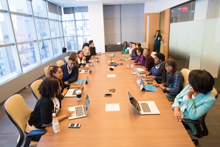 A modern conference room with a large wooden table that has several open laptops and people seated around it. The attendees, dressed in business attire, are engaged in a meeting. Natural light floods the room through large windows on one side, and a woman stands near the door at the back of the room.