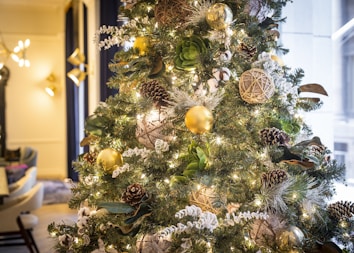 A beautifully decorated Christmas tree adorned with golden baubles, pinecones, and woven ornaments, surrounded by twinkling fairy lights. The greenery is lush and interspersed with hints of white and beige decorations, creating a festive and elegant look. In the background, soft lighting and modern furnishings add a cozy, warm atmosphere.