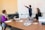 Three people are in a meeting room. One person is standing and pointing to a whiteboard filled with diagrams and flowcharts, while the other two are seated at a table with laptops and notebooks. The atmosphere appears focused and collaborative.