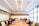 A group of people are seated around a long wooden conference table in a modern office room. The room has bright overhead lighting, large windows on one side, and a whiteboard on one wall. Each person is focused on their laptops or notes, suggesting a meeting or collaborative session.
