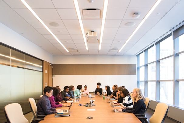 A group of people are seated around a long wooden conference table in a modern office room. The room has bright overhead lighting, large windows on one side, and a whiteboard on one wall. Each person is focused on their laptops or notes, suggesting a meeting or collaborative session.