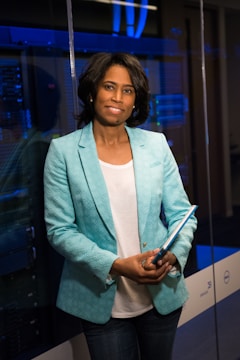 A person stands in a professional setting holding a blue folder. They are wearing a teal blazer over a white shirt and jeans. The background features glass walls and what appears to be a server room with blue lighting, suggesting a technological or business environment.