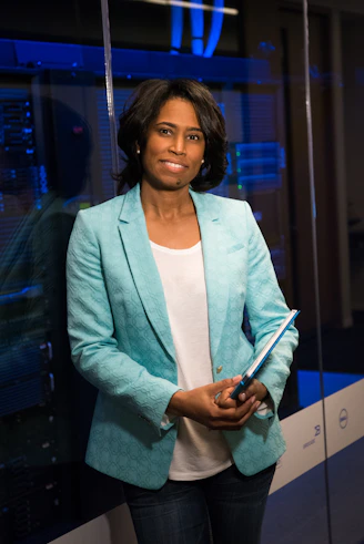 A person stands in a professional setting holding a blue folder. They are wearing a teal blazer over a white shirt and jeans. The background features glass walls and what appears to be a server room with blue lighting, suggesting a technological or business environment.