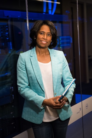 A person stands in a professional setting holding a blue folder. They are wearing a teal blazer over a white shirt and jeans. The background features glass walls and what appears to be a server room with blue lighting, suggesting a technological or business environment.