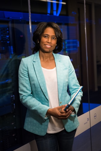 A person stands in a professional setting holding a blue folder. They are wearing a teal blazer over a white shirt and jeans. The background features glass walls and what appears to be a server room with blue lighting, suggesting a technological or business environment.